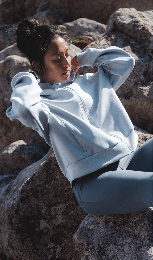 Athlete wearing the Jaida Full Zip Hooded Jacket in Frost Grey and Jaida Pocket Leggings in Granite Grey reclines on rocks in an outdoor setting.