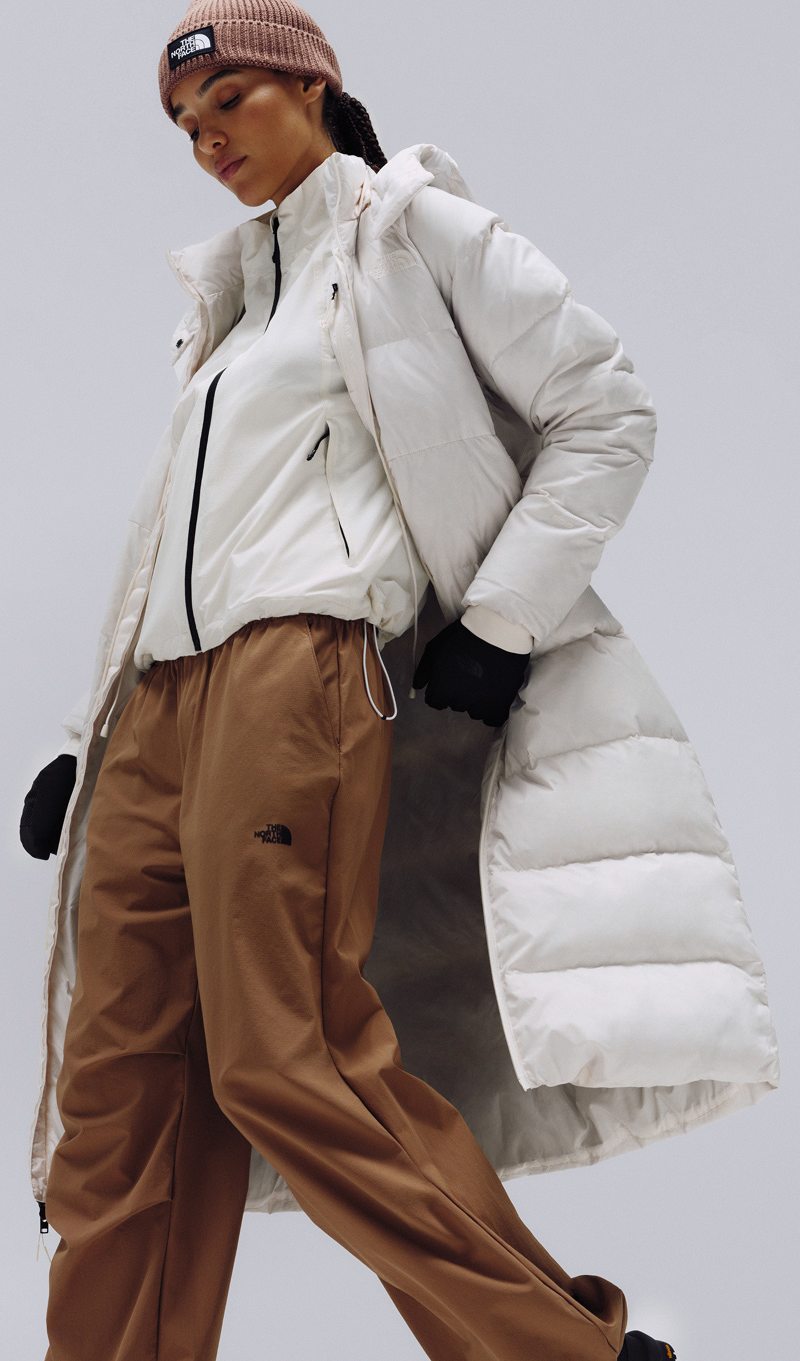 A female wearing The North Face Hydrenalite City Long Down Hooded Parka in White Dune and a brown knit beanie looks down against a neutral background