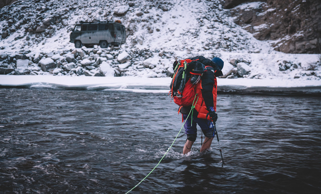 Jackson Marvell wading through an icy river with a fully loaded pack and his pant legs rolled up.