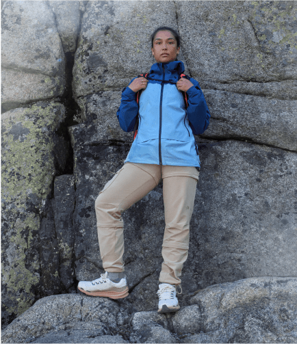 Woman standing in front of a rock wall wearing a blue jacket from The North Face and grasping straps from her backpack.