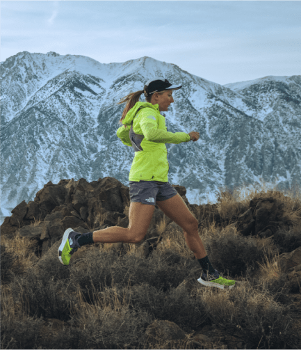 Woman running in a brigh yellow jacket in front of a snowy mountain range.