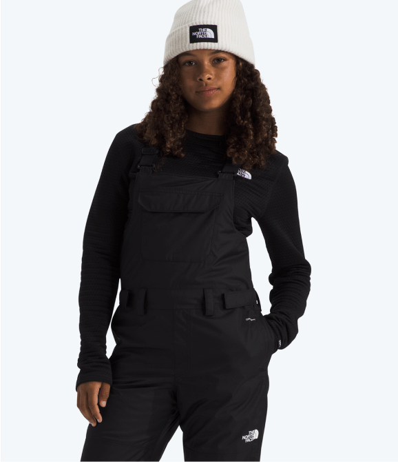 A child wearing black snow bibs from The North Face standing in front of a grey background.