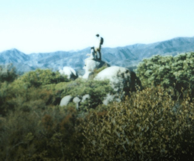 A panoramic shot of two hikers navigating over large boulders on rocky terrain.