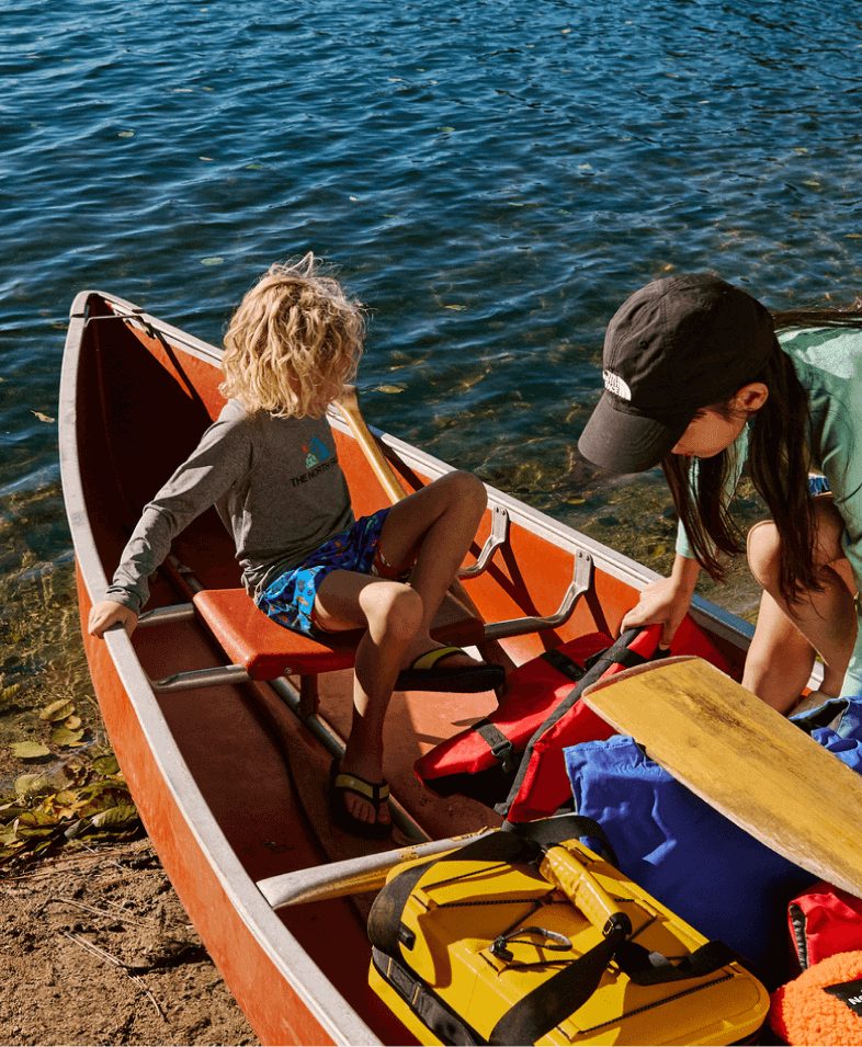 Two kids wearing gear from The North Face get into a canoe on the edge of a lake. 