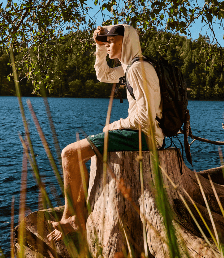 A man wearing sun-ready gear from The North Face sits on a tree stump while looking out at a lake.