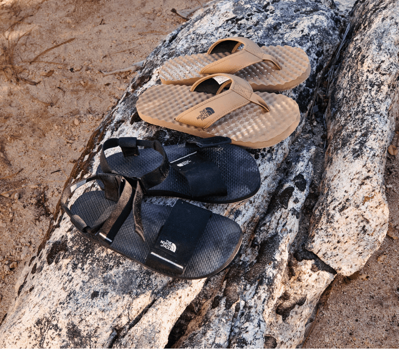 Two pairs of sandals from The North Face are set to the side on a sandy beach.