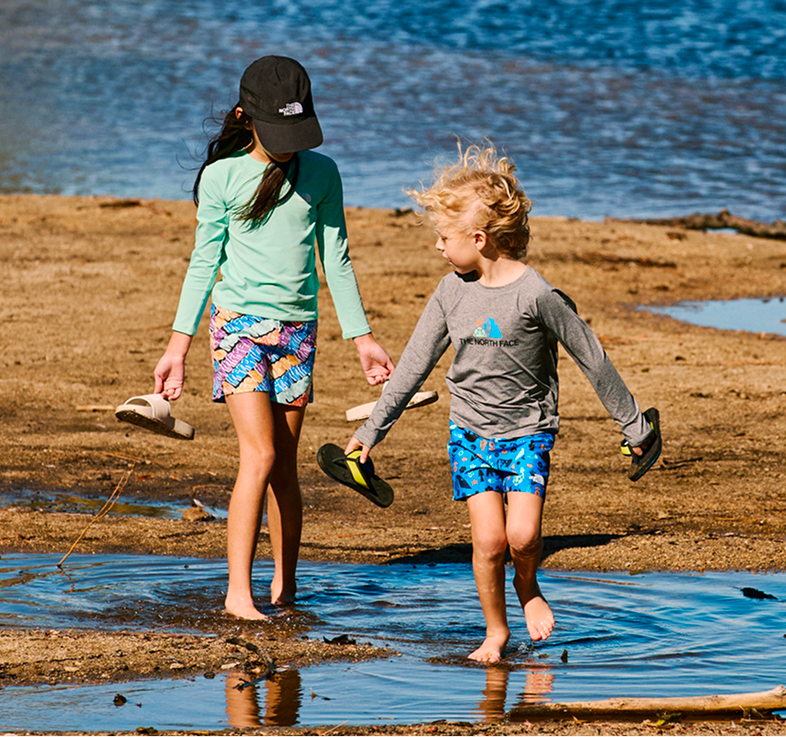 Two kids holding their sandals in their hands walk through water on a beach.