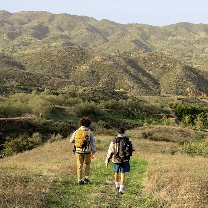 Two people walking in a field with mountains in the background, wearing backpacks from The North Face.