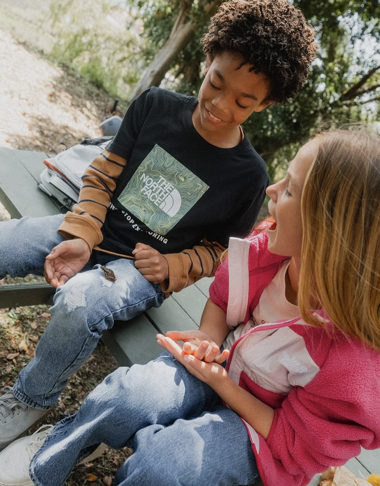 Two kids sitting on a picnic table looking at a small frog on the left kid’s leg.