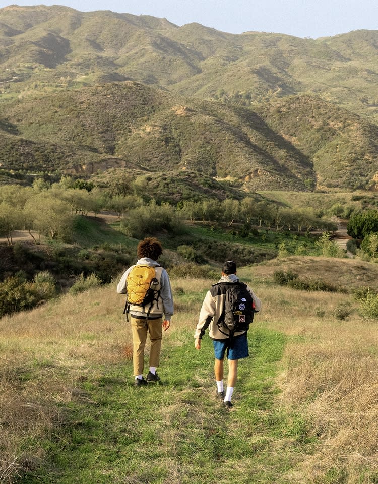Two kids walking through a field towards rolling hills. The one on the left has a yellow backpack, the one on the right has a black backpack with patches.