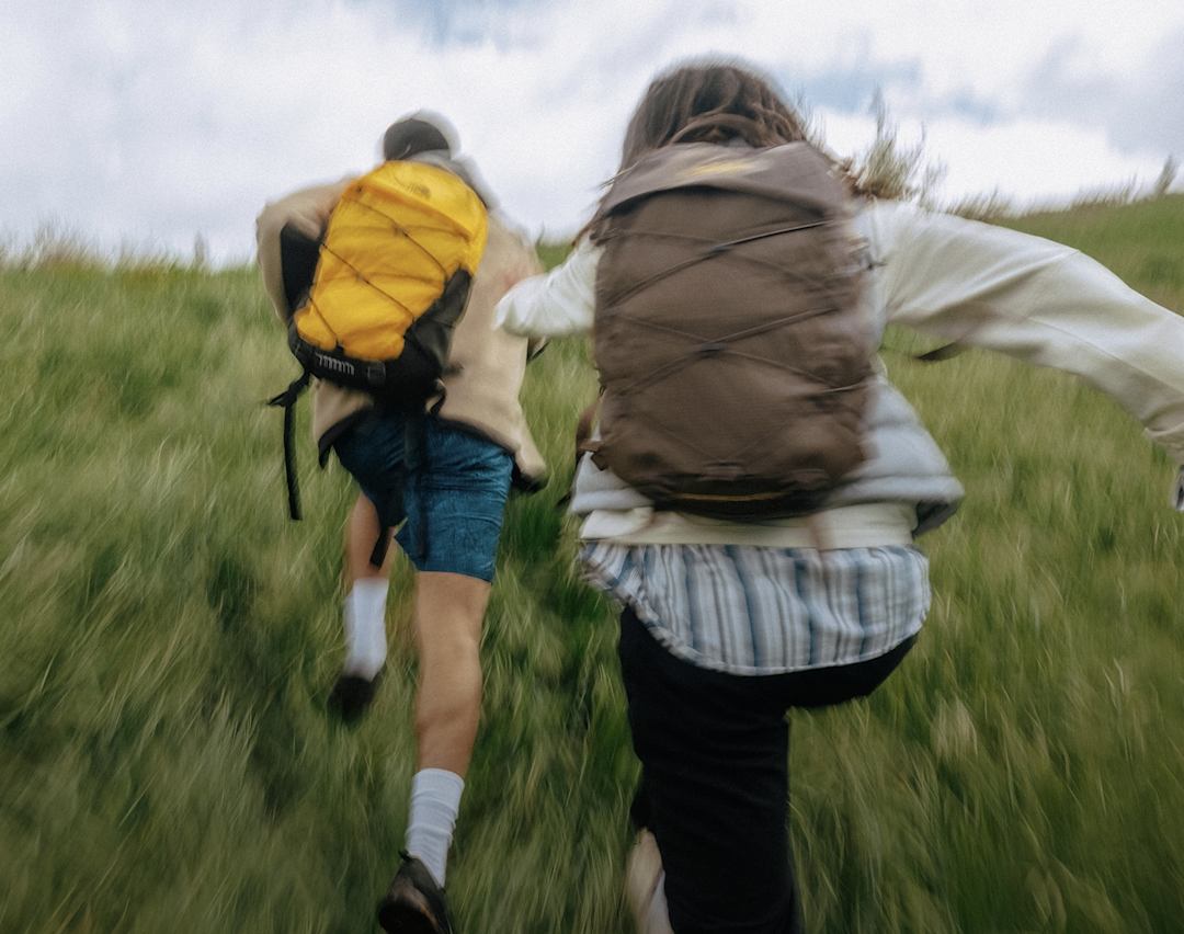 Two kids running through a field. The one on the left has a yellow backpack, the one on the right has a brown backpack.