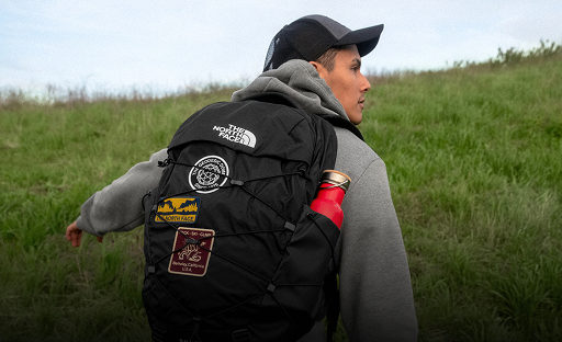A guy in a gray sweatshirt wearing a black backpack with patches. He’s walking through a field.