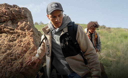 A guy in a taupe/black fleece walking up against a rock.