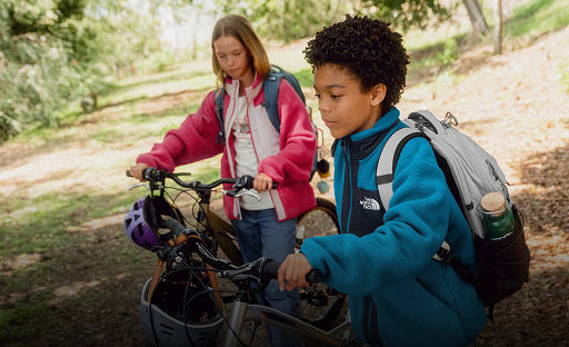 Two kids pushing bikes. The one in the foreground is in a blue jacket and the one in the background is in a pink jacket.