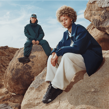 A woman in a blue jacket and white pants sitting on a rock in the foreground. In the background is a man sitting on a rock in an all green outfit.