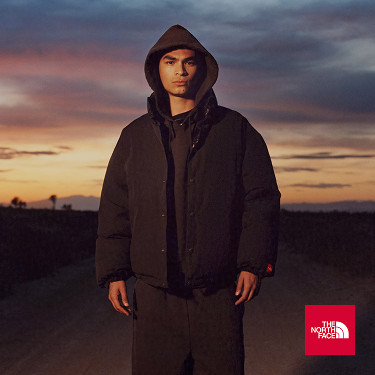 A man in a black jacket and black hoodie looking at the camera. The sun is setting in the background.