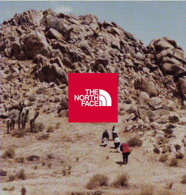 Five people walking out from a rocky hill. The woman to the left is in the foreground wearing a white sweatshirt with a red box label. There’s also a person in the background wearing a bouldering pad on their back.