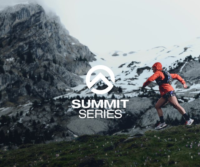A woman in a red jacket and floral vest running across grassy/rocky terrain. A the rocky cliffs in the background have snow on them.
