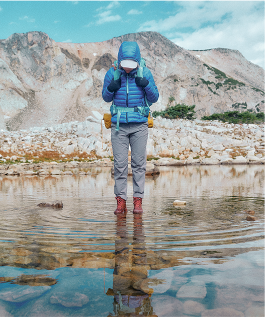 A hiker in The North Face gear stands in a shallow stream in front of a mountain range.