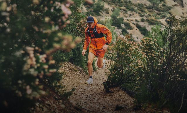 Athlete running up a dirt trail in an all-orange outfit with a black vest and running hat on.