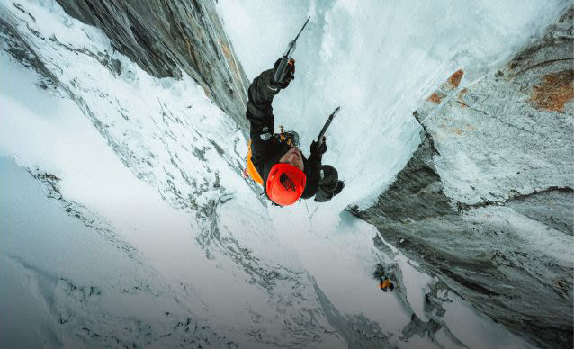 Wide shot of two alpinists tethered by a rope climbing up a snowy rock face.