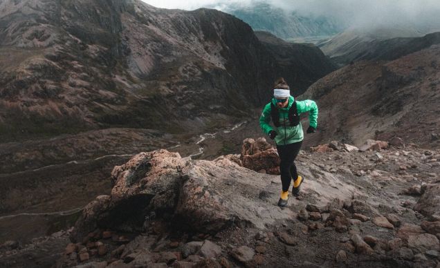 A woman in a white head band, turquoise jacket and black pants running up a rocky scramble.