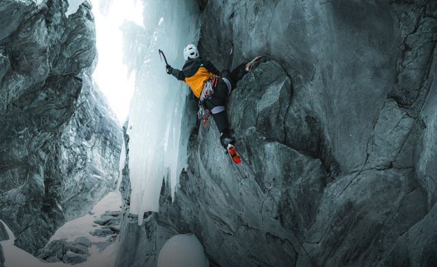 An ice climber in a yellow and black jacket climbing a rock face while sinking their left pick into a sheet of ice. They have a white helmet on.
