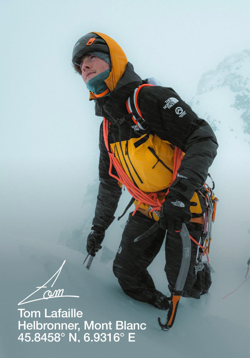 Tom in a yellow and black jacket trudging up a snow-covered hill with rope sashed across him. Someone is following behind. 