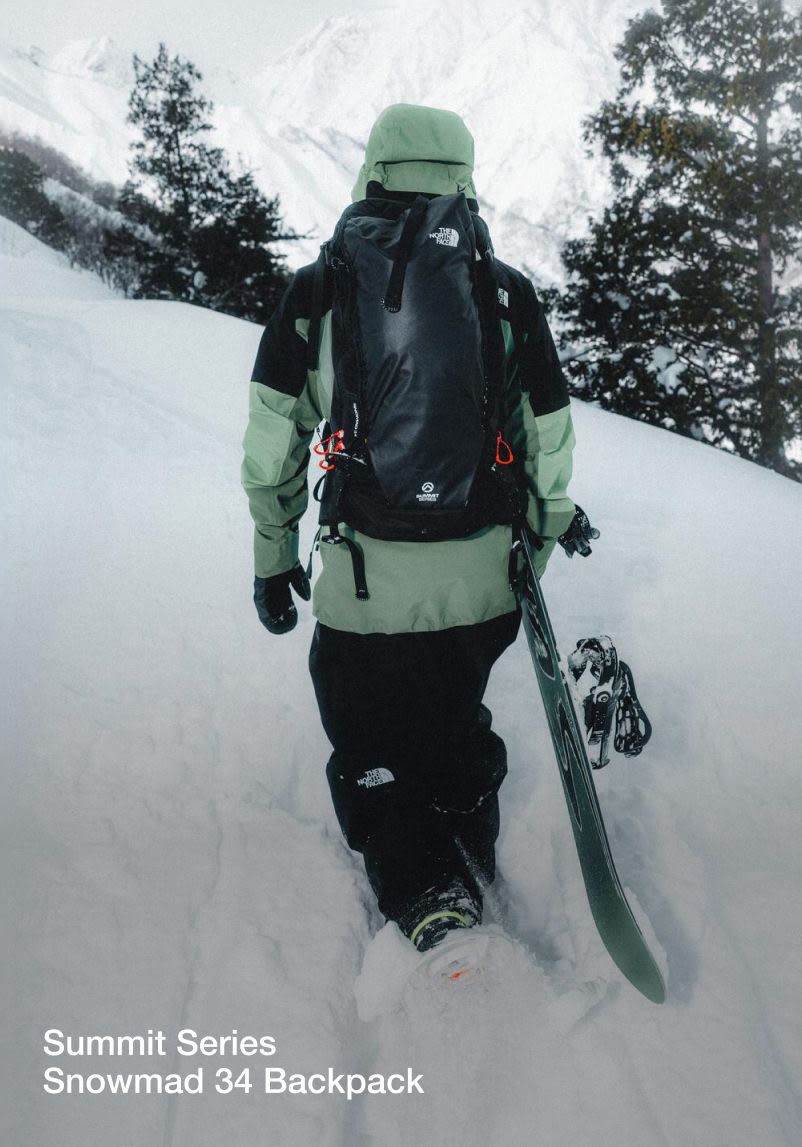 A shot of Nik from behind carrying his snowboard, wearing a green/black jacket with a black backpack. 