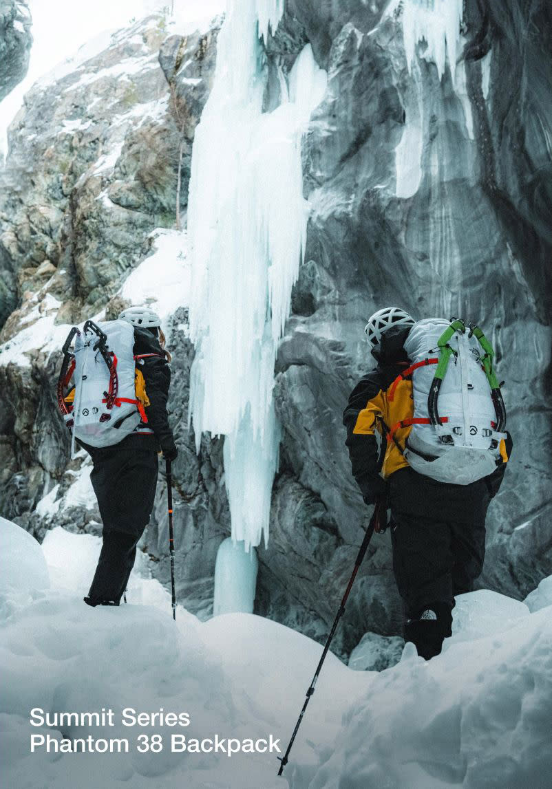 Two alpinists standing at the base of an ice wall with two white-ish/gray backpacks on.
