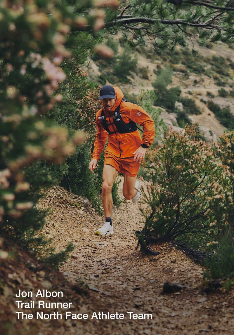 Jon running up a dirt trail in an all-orange outfit with a black vest and running hat on.