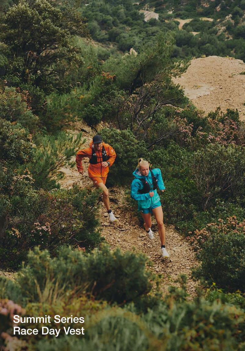 A wide shot of Jon running up hill in an all-orange outfit and black running vest. He’s following a woman in an all-blue outfit. 