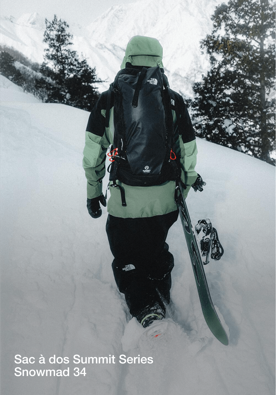 Une photo de Nik de dos portant son snowboard, vêtu d'une veste verte/noire avec un sac à dos noir.