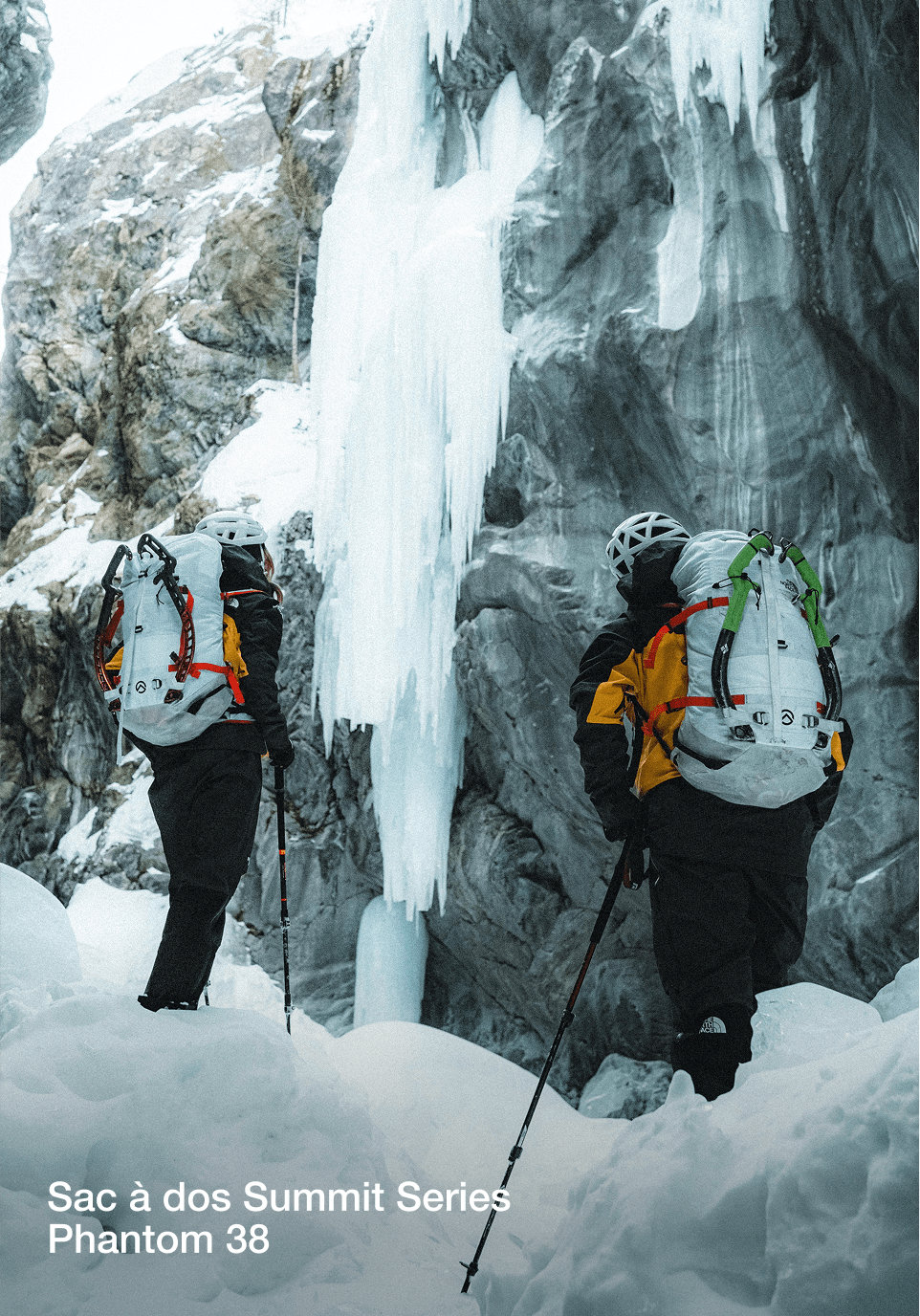 Deux alpinistes se tiennent au pied d'une paroi de glace, deux sacs à dos blanchâtres/gris sur le dos. Ils tournent le dos à la caméra.