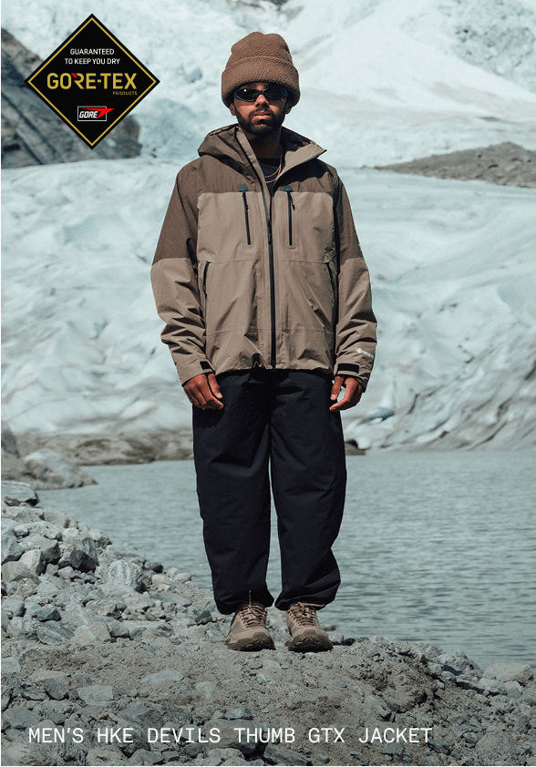 A man in a brown beanie and brown jacket looking directly at the camera with sunglasses on. He’s in front of a glacier.