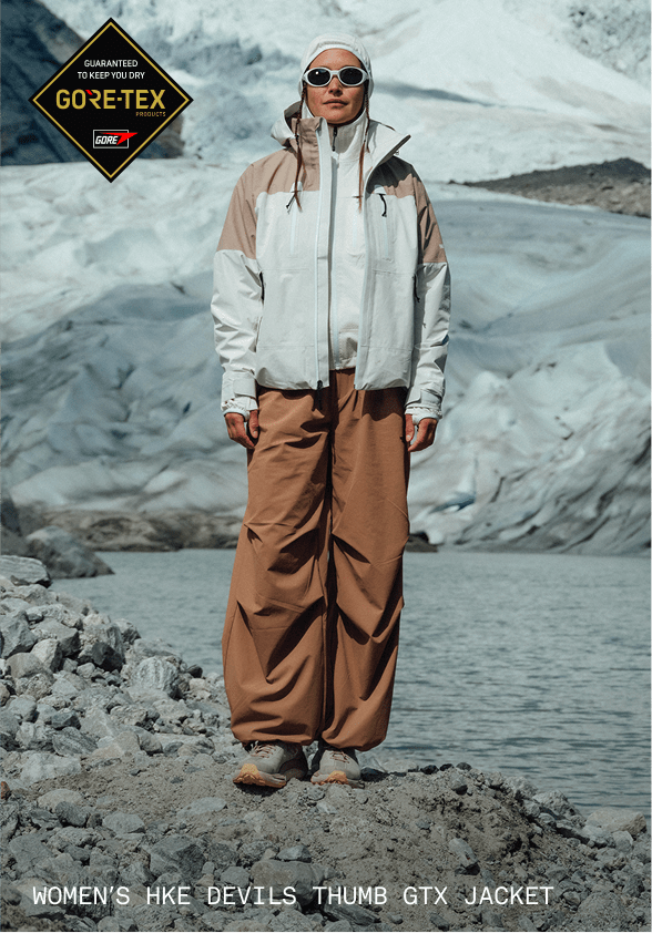 A woman in a white jacket and white hoodie looking directly at the camera with sunglasses on. She’s in front of a glacier. 