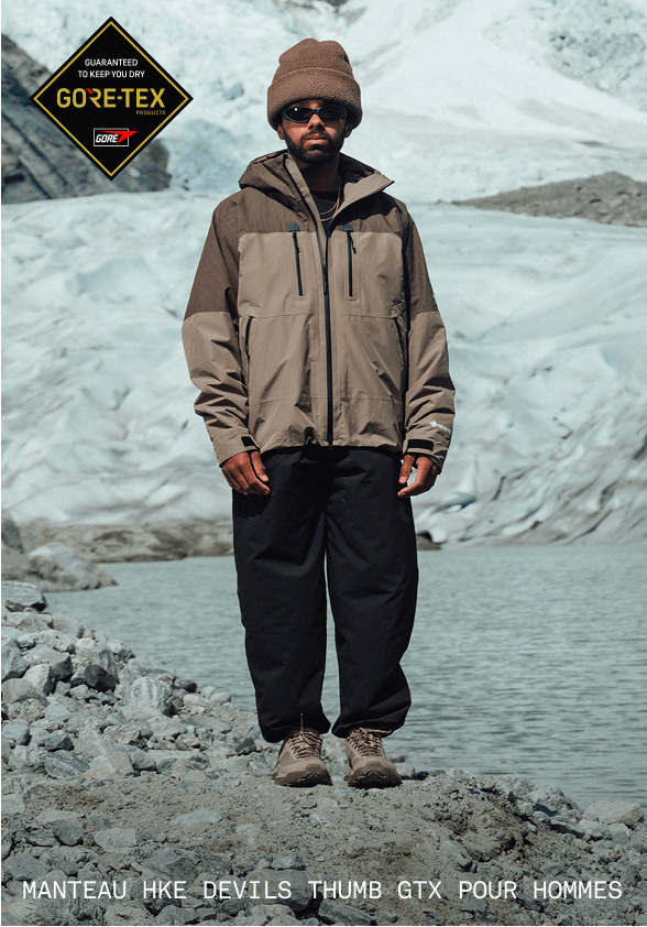 Un homme vêtu d’une tuque et d’un manteau brun et portant des lunettes de soleil regardant directement la caméra. Il se tient devant un glacier.