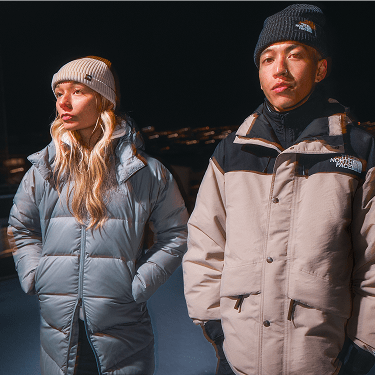 Two people stand outdoors at night wearing The North Face winter jackets and knit beanies, with city lights glowing behind them.