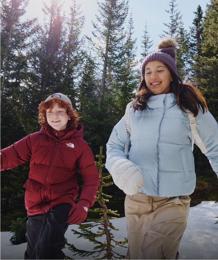 A boy in a red jacket and a girl in a light blue jacket running with pine trees behind them.