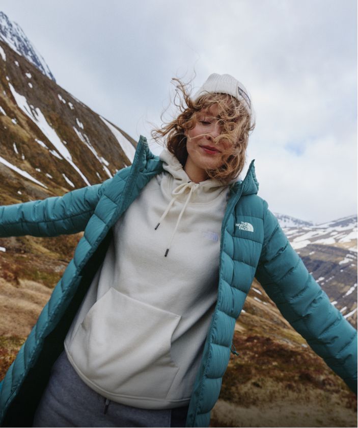 A woman in a white hoodie/white beanie/turquoise jacket with her arms stretched out. 