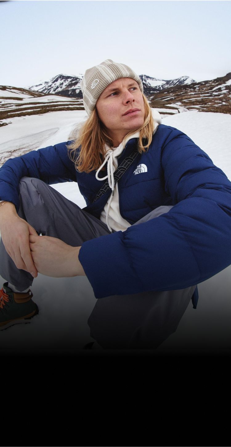 A guy in a white hoodie/tan beanie/navy jacket sitting in the snow. He’s looking away from the camera.