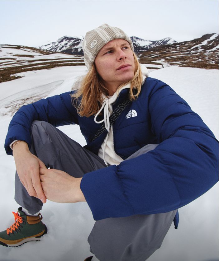 A guy in a white hoodie/tan beanie/navy jacket sitting in the snow. He’s looking away from the camera. 