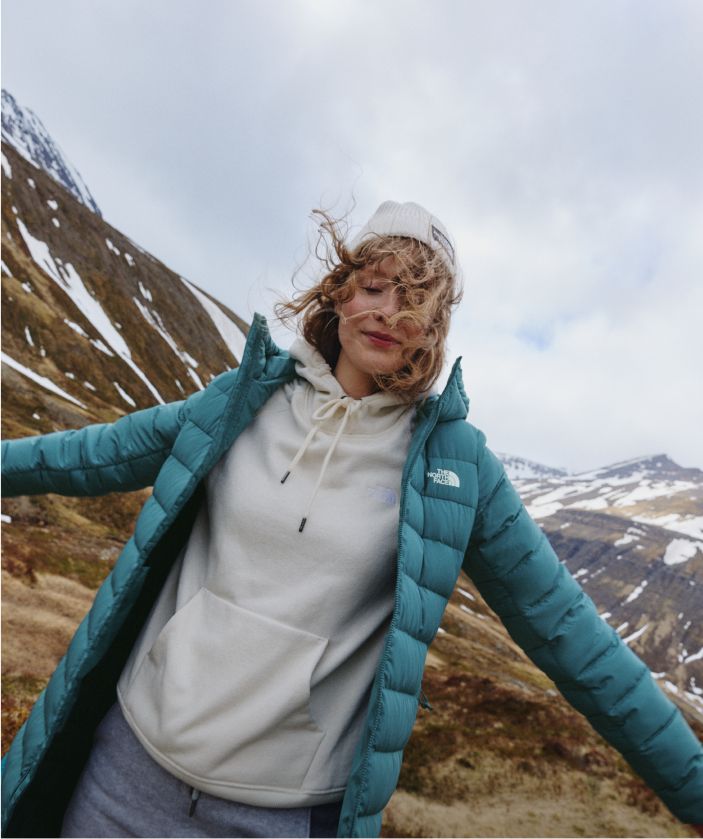 A woman in a white hoodie/white beanie/turquoise jacket with her arms stretched out. 