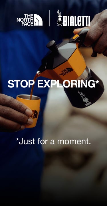 Person wearing a blue jacket holding a yellow TNF X Bialetti Moka Express coffee maker in one hand and a yellow TNF branded cup in the other. The coffee is actually puring out of the coffee maker’s spout.