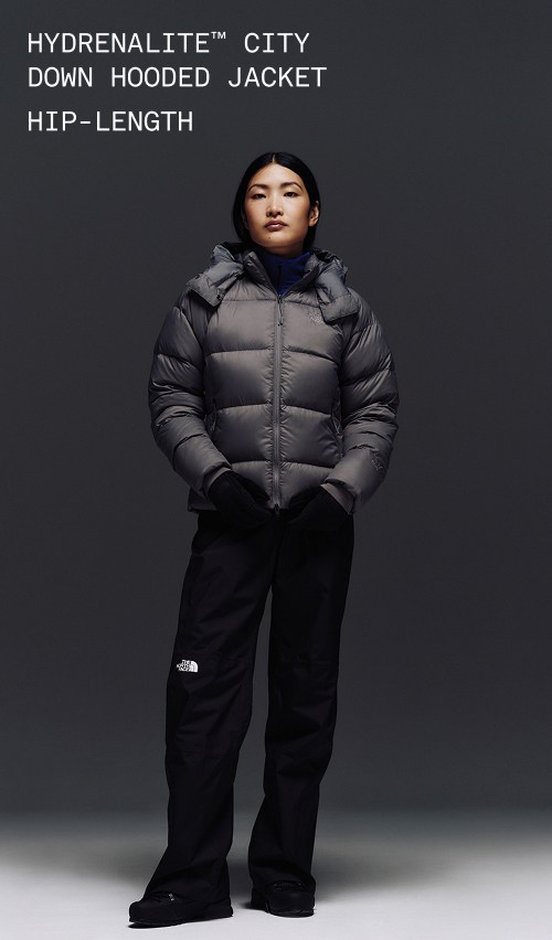 A female wearing The North Face Hydrenalite City Long Down Hooded Parka in White Dune and a brown knit beanie looks down against a neutral background. 