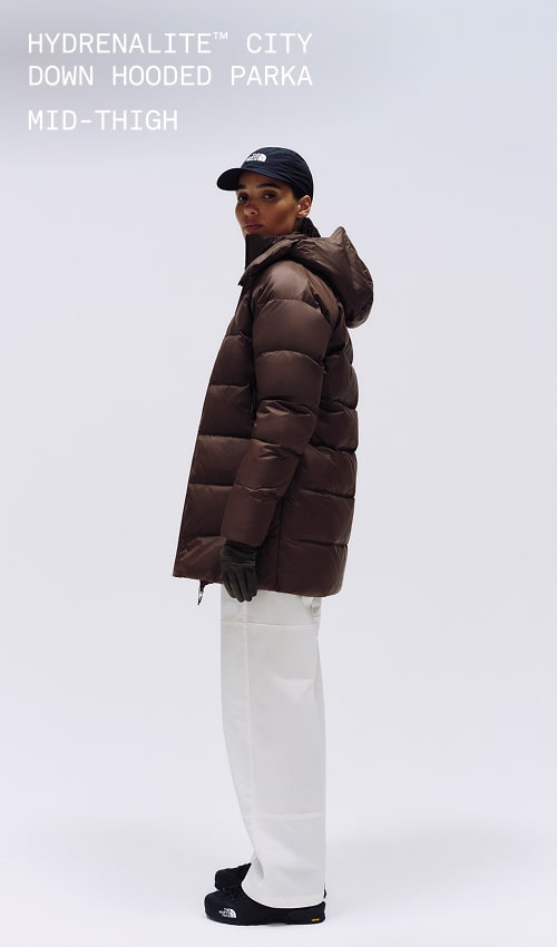 A female wearing The North Face Hydrenalite City Long Down Hooded Parka in White Dune and a brown knit beanie looks down against a neutral background. 