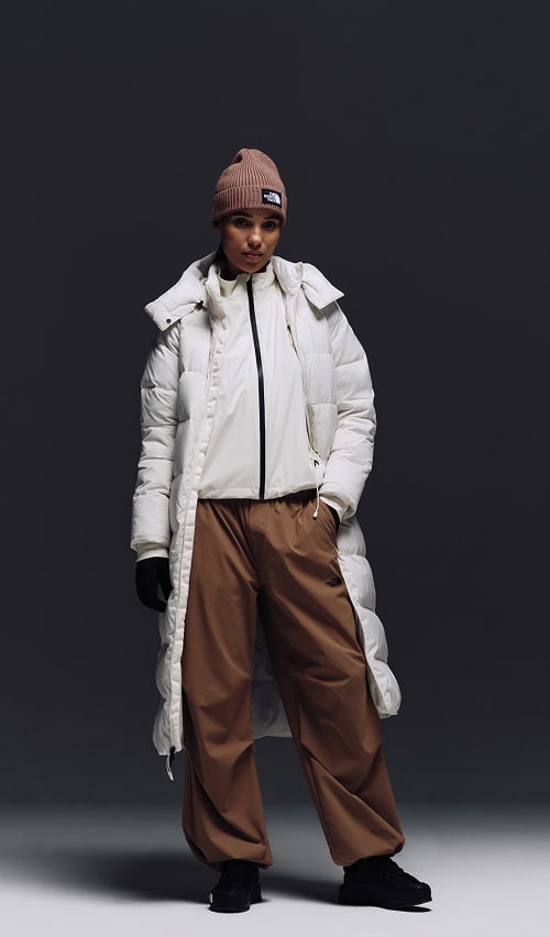 A female wearing The North Face Hydrenalite City Long Down Hooded Parka in White Dune and a brown knit beanie looks down against a neutral background. 