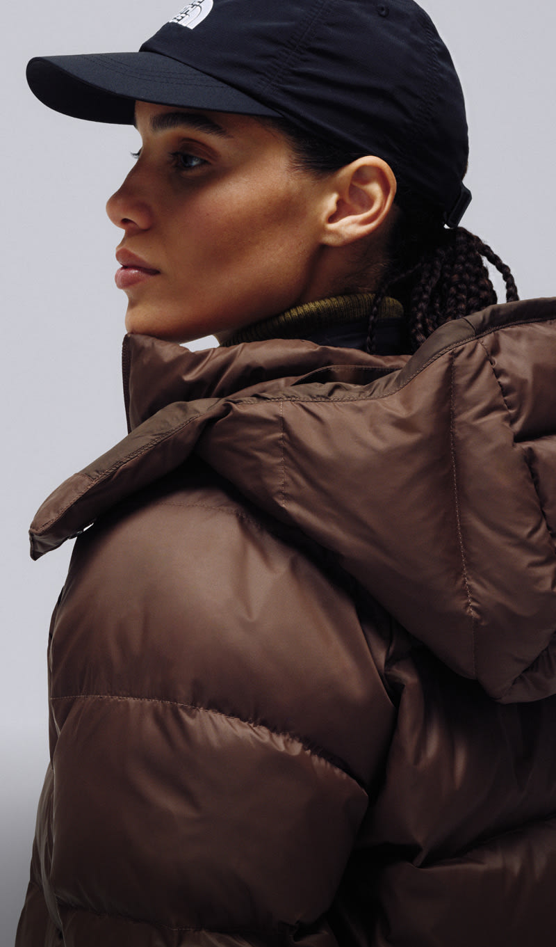 A female wearing The North Face Hydrenalite City Long Down Hooded Parka in White Dune and a brown knit beanie looks down against a neutral background. 