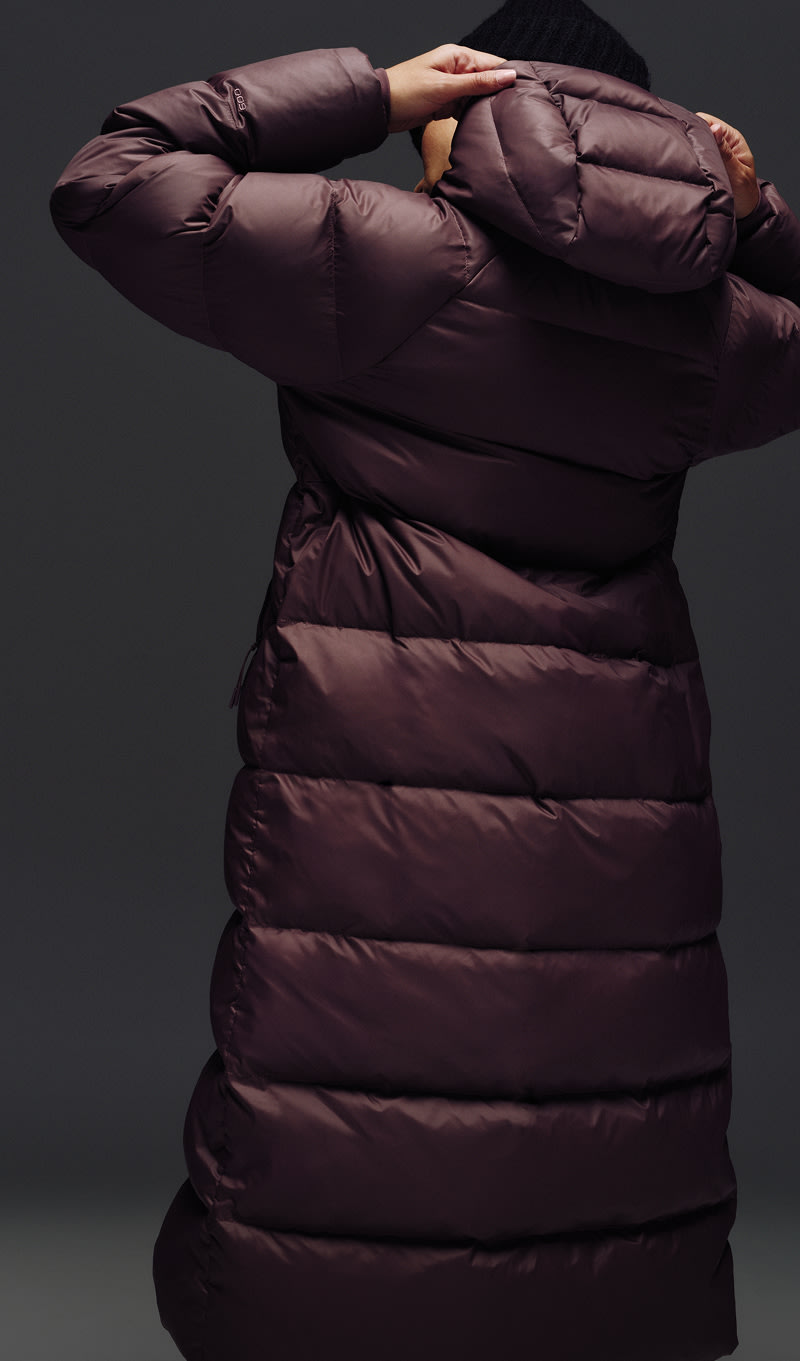 A female wearing The North Face Hydrenalite City Long Down Hooded Parka in White Dune and a brown knit beanie looks down against a neutral background. 