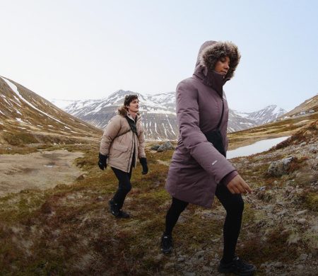 A person in a purple jacket with the hood up walking in front of someone in a tan jacket. They’re headed uphill. 